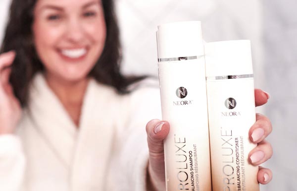 Lifestyle shot of a woman in a white robe holding up the ProLuxe Shampoo and Conditioner bottles in one hand.